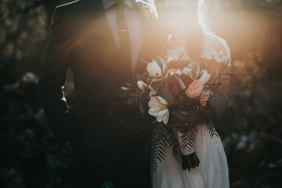 Low sun flares behind a couple holding a loose bouquet of white roses and fern.