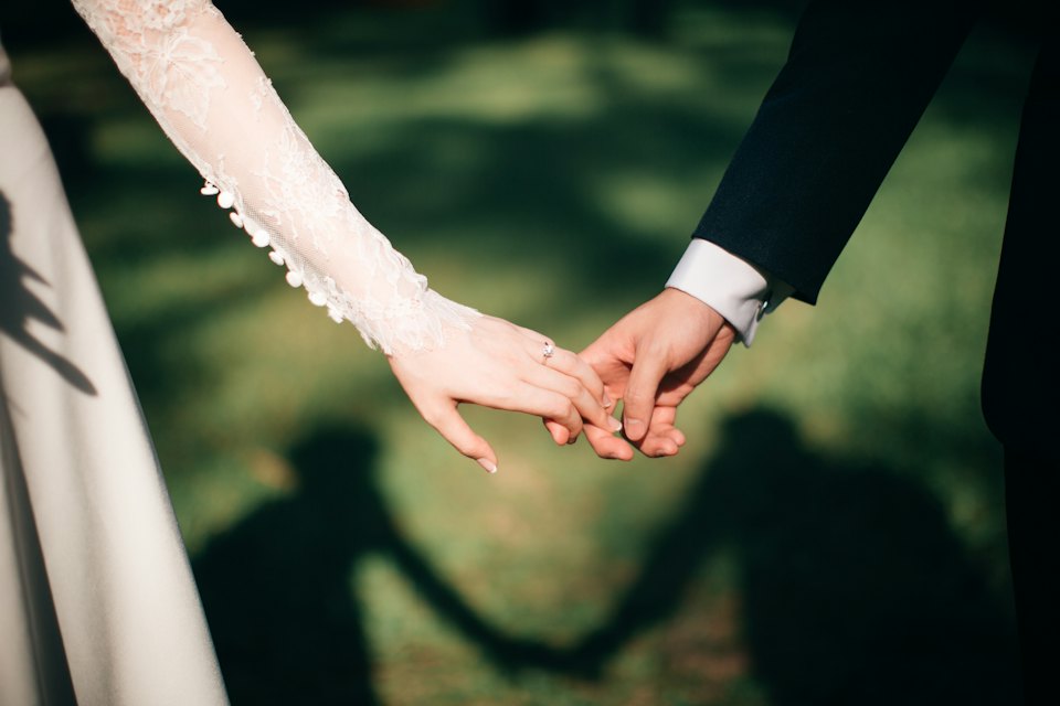Close crop of a bride in a lace sleeve and a groom holding hands, shadows across the lawn.