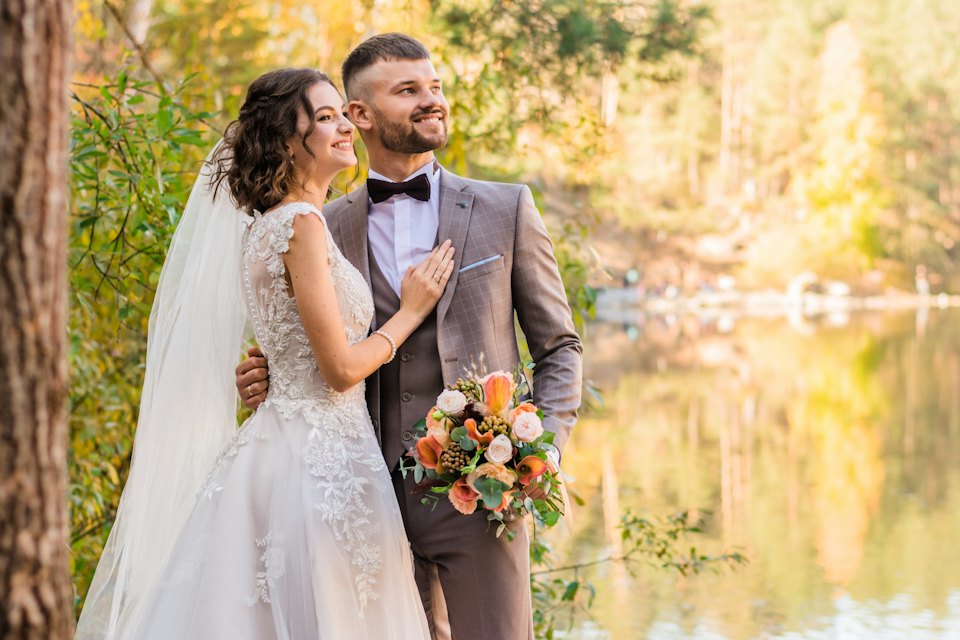 A smiling couple stand by a still lake framed by autumn trees, holding coral roses.