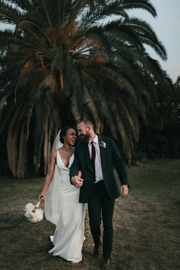 A laughing bride in a silk gown and a groom in a dark green suit beneath a towering palm at dusk.