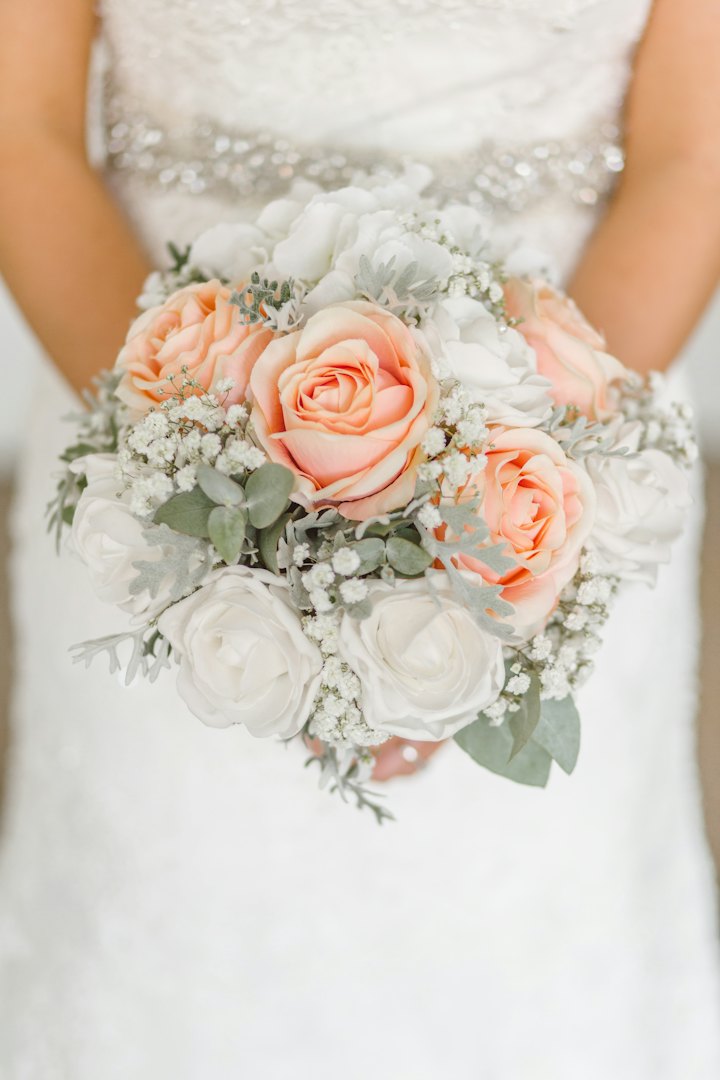 A bride holds a round bouquet of peach and ivory roses against a beaded gown.