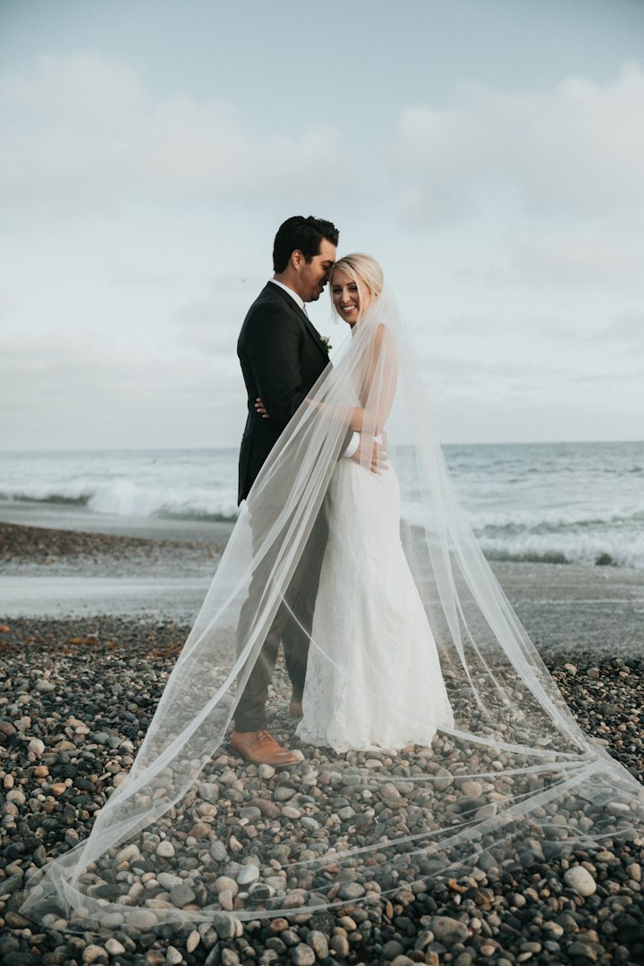 A couple embrace on a pebble beach, a cathedral-length veil spreading across the stones.